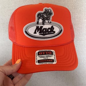 May include: Orange trucker hat with a black and white bulldog graphic and the name "Mack". The hat has a mesh back and a curved brim. The text "World Famous Foam Front" is also visible.