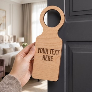 Puede incluir: Un colgador de puerta de madera de color claro con un recorte circular en la parte superior y un cuerpo rectangular. El texto "YOUR TEXT HERE" está grabado en una fuente oscura sin remates en la superficie frontal de la madera, colgado en una puerta gris oscuro.