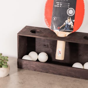 May include: A dark brown wooden table tennis paddle rack with circular cutouts on the top surface. The rack holds a red and black table tennis paddle with a light wood handle and several white table tennis balls inside the rectangular base.