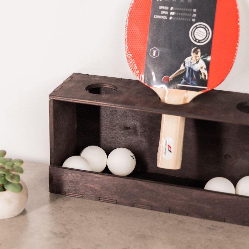 May include: A dark brown wooden table tennis paddle rack with circular cutouts on the top surface. The rack holds a red and black table tennis paddle with a light wood handle and several white table tennis balls inside the rectangular base.
