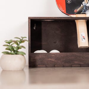 May include: A dark brown wooden wall-mounted storage box featuring a rectangular frame with visible joinery. Inside the box are three white ping pong balls. A table tennis paddle with a red and black face is inserted into the top right corner.