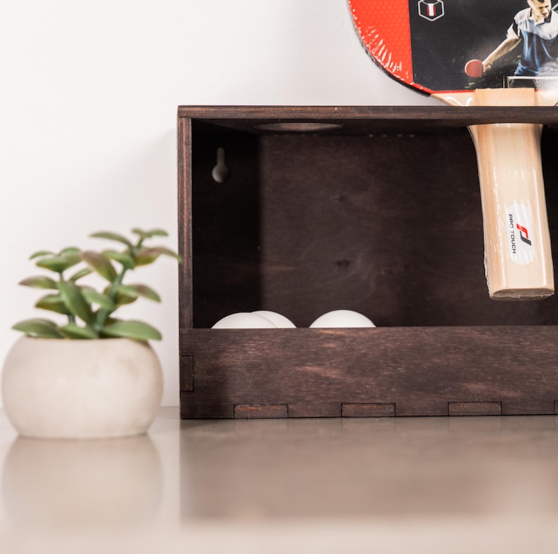 May include: A dark brown wooden wall-mounted storage box featuring a rectangular frame with visible joinery. Inside the box are three white ping pong balls. A table tennis paddle with a red and black face is inserted into the top right corner.