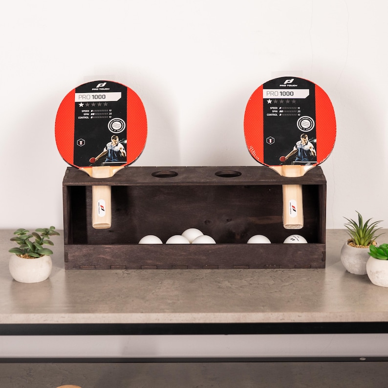 May include: A dark brown wooden storage rack holding two Pro 1000 table tennis paddles with red and black faces, and five white ping pong balls resting inside the base. The paddles feature performance ratings and a graphic of a player.