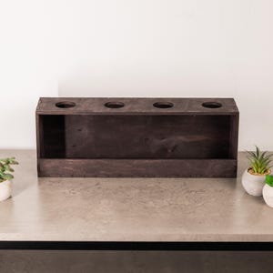 May include: A rectangular dark brown wooden box featuring four circular cutouts along the top panel. The box has a flat front face and visible joinery details along the edges, presenting a simple, functional design for holding items.