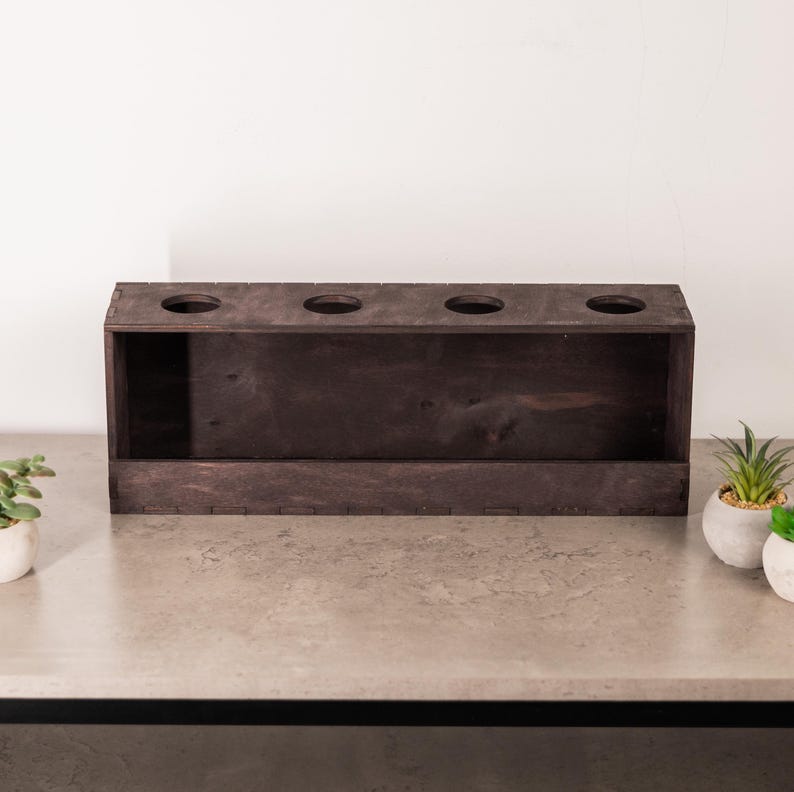 May include: A rectangular dark brown wooden box featuring four circular cutouts along the top panel. The box has a flat front face and visible joinery details along the edges, presenting a simple, functional design for holding items.