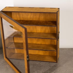 May include: A wooden display case with a glass door, featuring multiple shelves. The case is made of light brown wood with a darker brown trim. The door is open, revealing the interior shelves. The case is designed to showcase collectibles or small items.