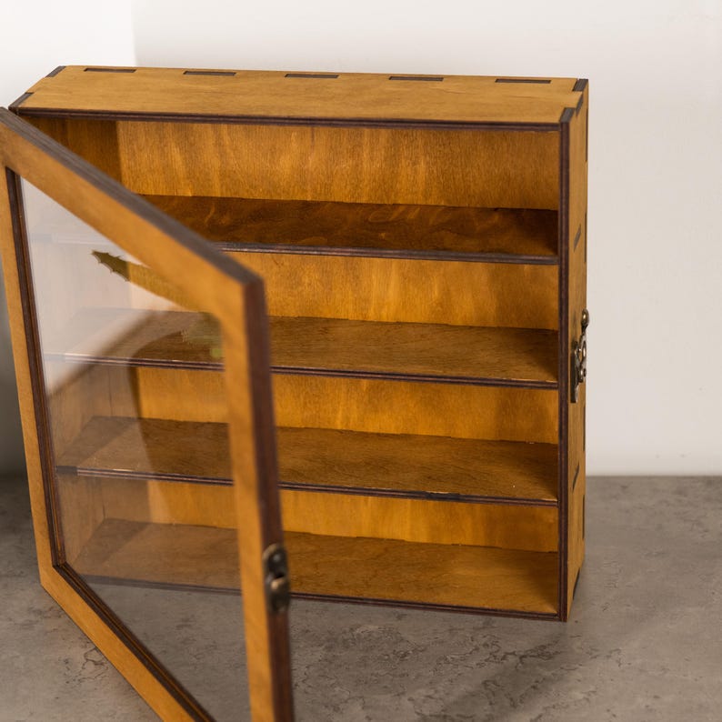 May include: A wooden display case with a glass door, featuring multiple shelves. The case is made of light brown wood with a darker brown trim. The door is open, revealing the interior shelves. The case is designed to showcase collectibles or small items.