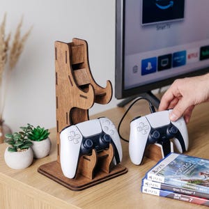 Puede incluir: Un soporte de madera para mandos de juegos con dos mandos blancos y negros. El soporte es de madera marrón claro y tiene un diseño único. Se ven dos cajas de videojuegos en primer plano y un televisor al fondo.