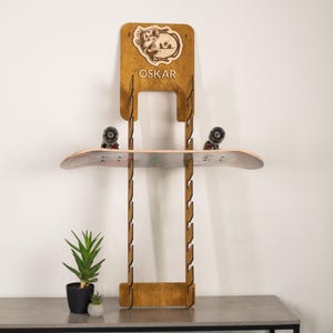 May include: A vertical wooden skateboard wall rack with a dark brown finish, featuring a laser-engraved fish graphic and the name OSKAR at the top. The rack has multiple side notches for storage and holds a single skateboard horizontally.