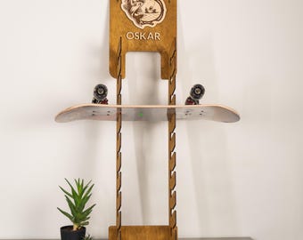 Puede incluir: Un soporte de pared vertical de madera para monopatines con acabado marrón oscuro, que presenta un gráfico de pez grabado con láser y el nombre OSKAR en la parte superior. El soporte tiene varias muescas laterales y sostiene un monopatín.