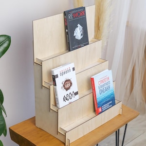 May include: A wooden, tiered bookshelf displaying three books with colorful covers. The bookshelf is made of light-colored wood and has a stepped design. The books are arranged on different levels, showcasing their titles and cover art.