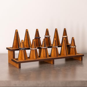 May include: A wooden ring display stand with multiple cone-shaped holders. The stand is made of light brown wood and holds several rings of various designs. The rings are silver and gold, with some featuring gemstones. The stand is on a gray surface.