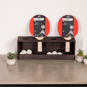 Könnte beinhalten: Ein dunkelbrauner Holz-Tischtennisschlägerhalter mit zwei roten und schwarzen PRO 1000 Schlägern und weißen Tischtennisbällen. Der Halter hat zwei Löcher für die Schläger und ein unteres Fach für die Bälle.