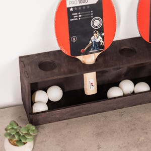 May include: A dark brown wooden table tennis paddle rack with circular cutouts for two paddles. The rack features a lower storage compartment containing five white ping pong balls. One paddle is inserted, displaying the text PRO 1000.