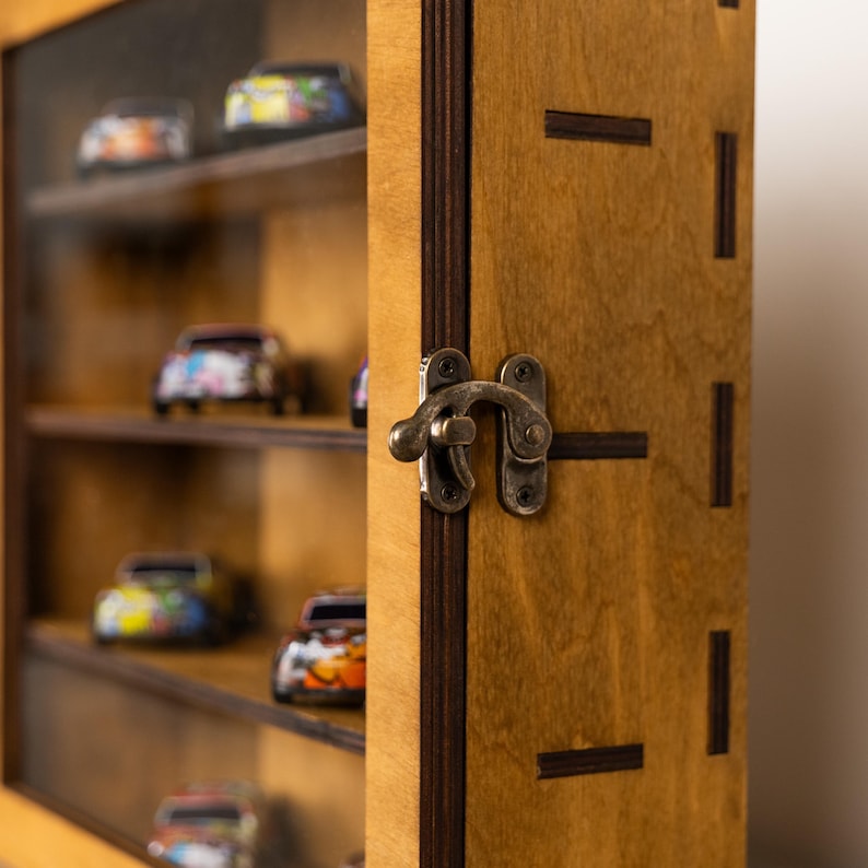 May include: A wooden display case with a glass front, showcasing several miniature toy cars on shelves. The case has a warm, natural wood finish and a metal latch. The toy cars are brightly colored.