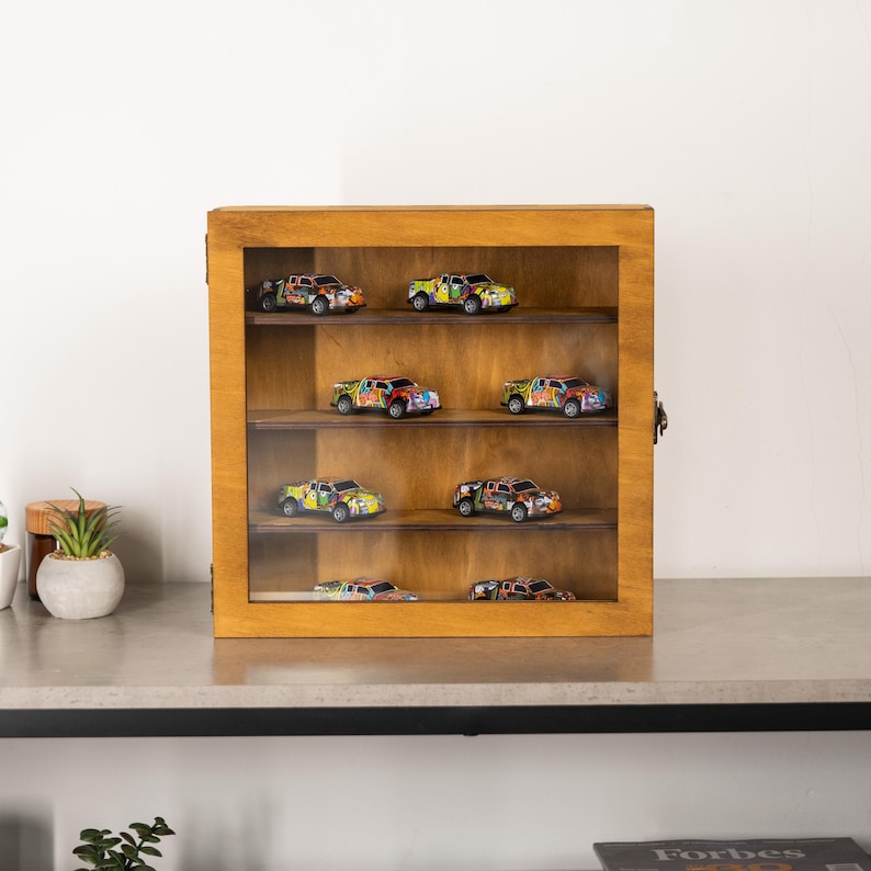 May include: A wooden display case with a glass front, showcasing several colorful toy trucks. The trucks are painted with a variety of colors and designs. The case has multiple shelves and a dark metal clasp.
