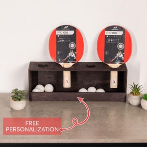 May include: A dark brown wooden table tennis rack with two slots holding Pro Touch 1000 paddles and a lower compartment containing five white ping pong balls. A red rectangular graphic with white text reads FREE PERSONALIZATION.