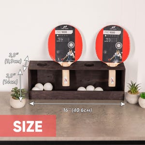 May include: A dark brown wooden table tennis rack holding two Pro 1000 paddles and several white balls. The rack measures 16 inches wide, 3.9 inches deep, and 5.5 inches high. The paddles feature red rubber surfaces and light wood handles.