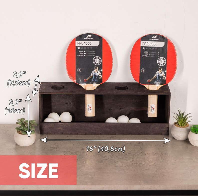 May include: A dark brown wooden table tennis rack holding two Pro 1000 paddles and several white balls. The rack measures 16 inches wide, 3.9 inches deep, and 5.5 inches high. The paddles feature red rubber surfaces and light wood handles.