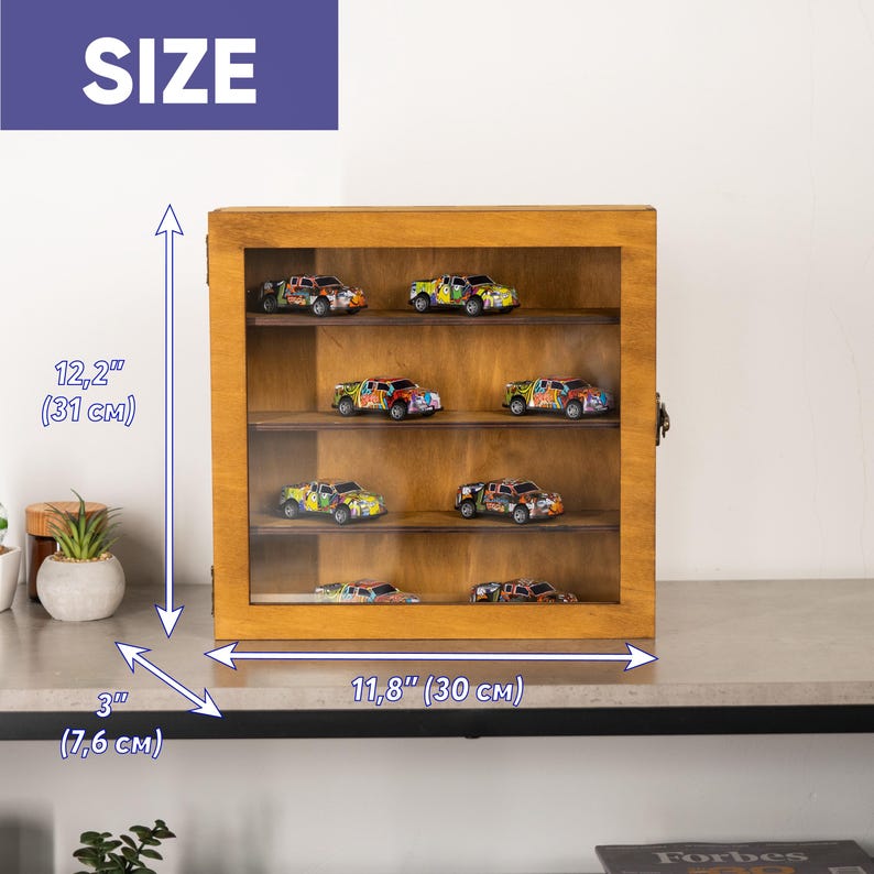 May include: A wooden display case with a glass front, holding several colorful toy trucks. The case is 12.2 inches tall, 11.8 inches wide, and 3 inches deep. The trucks are painted with various designs and colors.