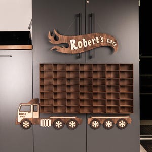 May include: A wooden car display shelf shaped like a truck, featuring the text "Robert's truck" and "Robert's cars". The shelf is designed with numerous small compartments for toy cars. The design incorporates decorative wheels and a flame-shaped sign.