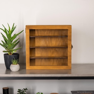 May include: A wooden display case with three shelves. The case is made of light brown wood and has a hinged door. The case is empty and ready to be filled with collectibles. The case is sitting on a gray surface.