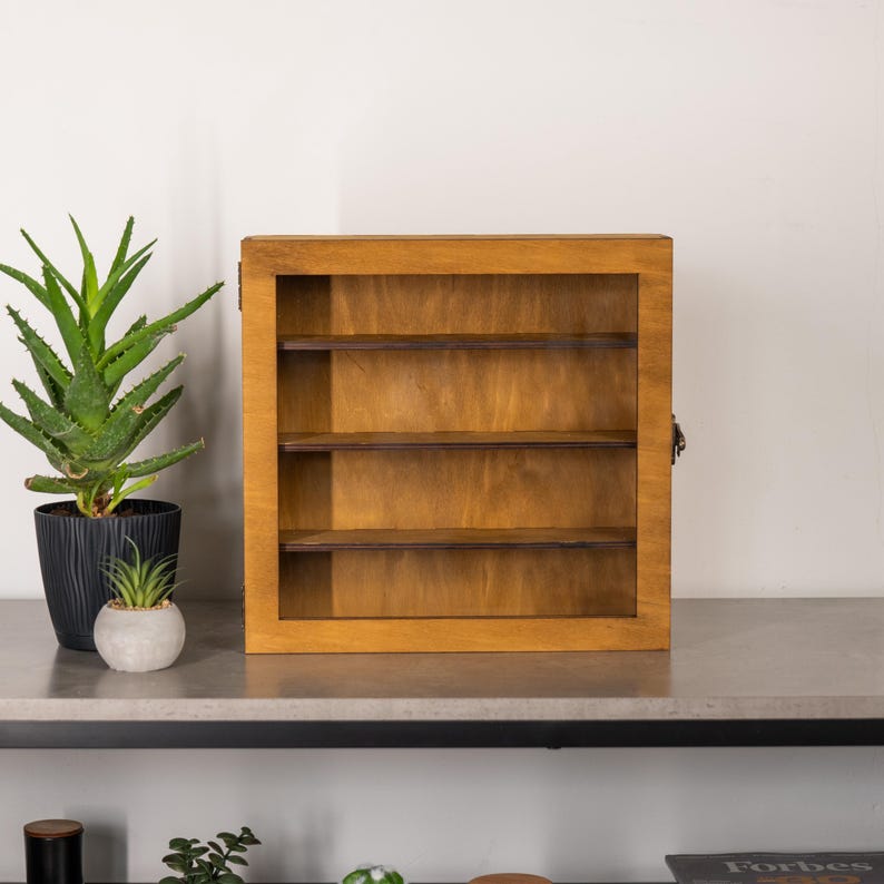 May include: A wooden display case with three shelves. The case is made of light brown wood and has a hinged door. The case is empty and ready to be filled with collectibles. The case is sitting on a gray surface.