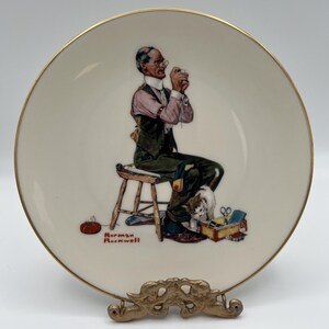 May include: A decorative plate featuring a seated man with glasses, sewing. The man is wearing a vest and patched pants. A small dog and sewing kit are at his feet. The plate has a gold rim and the artist's signature.