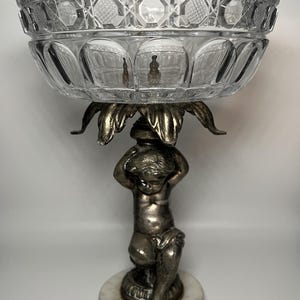 May include: A clear glass bowl with a decorative cut-glass pattern, supported by a silver-coloured metal base featuring a cherub figure. The base rests on a round, white marble pedestal. A classic decorative piece.