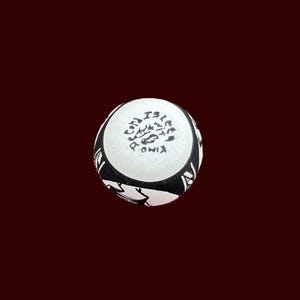 May include: A round, white ceramic object with black decorative patterns and text. The top surface features a floral design and text. The object is set against a dark red background.