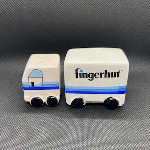May include: Two white ceramic salt and pepper shakers shaped like small trucks. One has a house design, the other has the word "fingerhut" in black. Both have blue stripes and black wheels.
