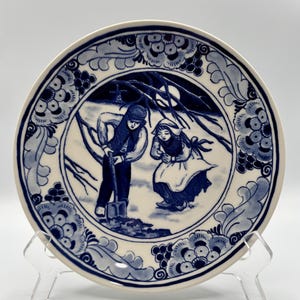 May include: A decorative ceramic plate with a blue and white illustration of two figures in a winter scene. The plate has a floral border and is displayed on a clear stand. The figures are dressed in traditional clothing.
