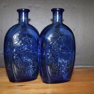 Puede incluir: Dos botellas de vidrio azul cobalto con cuerpos ovalados y cuellos largos. Cada botella presenta un diseño en relieve detallado. Las botellas se exhiben sobre una superficie de madera contra un fondo oscuro.