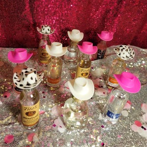 May include: Miniature cowboy hats in white, pink, and cow print atop small liquor bottles. The bottles are arranged on a sequined surface with a pink sequined backdrop. The bottles have labels with the brand name "Tito's" and other brands.