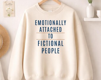 May include: Cream-colored sweatshirt with the text "EMOTIONALLY ATTACHED TO FICTIONAL PEOPLE" in navy blue. The sweatshirt is hanging on a wooden hanger against a white wall. A small "hello" sign and a vase of flowers are visible.