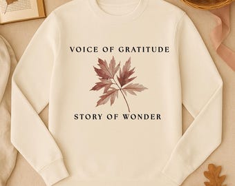 Puede incluir: Sudadera de color crema con el texto "VOICE OF GRATITUDE" y "STORY OF WONDER" en negro. Una ilustración detallada de una hoja de color marrón rojizo está centrada debajo del texto. La sudadera forma parte de una colección con temática otoñal.