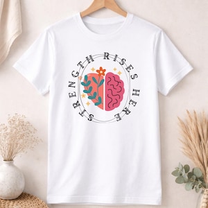 Peut inclure: T-shirt blanc avec un motif circulaire représentant un cœur et un cerveau. Le motif est divisé en deux, avec un cœur rose et un cerveau rose. Les mots "STRENGTH RISES HERE" sont imprimés autour du motif.
