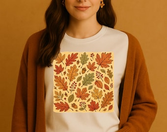 Puede incluir: Una mujer con una camiseta blanca con un diseño cuadrado de hojas de otoño en tonos rojos, naranjas, amarillos y verdes. También lleva una rebeca marrón y pantalones verde oscuro. El fondo es de color marrón liso.