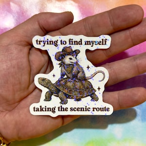 May include: A die-cut sticker featuring a brown opossum wearing a cowboy hat and bandana, riding on the back of a brown turtle. The text "trying to find myself" appears above and "taking the scenic route" below, with a holographic glitter finish.