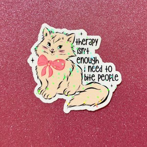 May include: A sticker featuring a cartoon cat with a pink bow, and the text "therapy isn't enough; I need to bite people." The cat is light brown with green and yellow accents, set against a pink glitter background.
