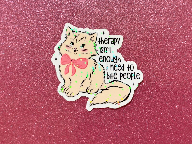 May include: A sticker featuring a cartoon cat with a pink bow, and the text "therapy isn't enough; I need to bite people." The cat is light brown with green and yellow accents, set against a pink glitter background.