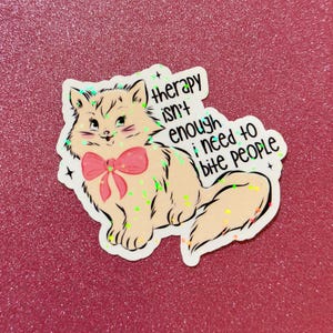 May include: A sticker featuring a cartoon cat with a pink bow. The cat is light peach with black outlines and green accents. Text on the sticker reads "therapy isn't enough, I need to bite PEOPLE."