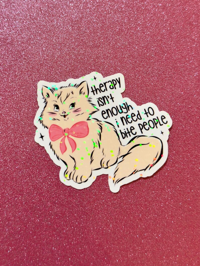 May include: A sticker featuring a cartoon cat with a pink bow. The cat is light peach with black outlines and green accents. Text on the sticker reads "therapy isn't enough, I need to bite PEOPLE."