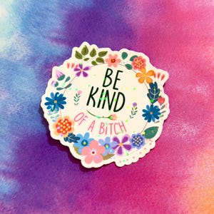 May include: A white sticker with a floral wreath design and the text "BE KIND OF A BITCH". The sticker features colorful flowers in shades of blue, pink, orange, and purple, with green leaves. The background is a tie-dye pattern.