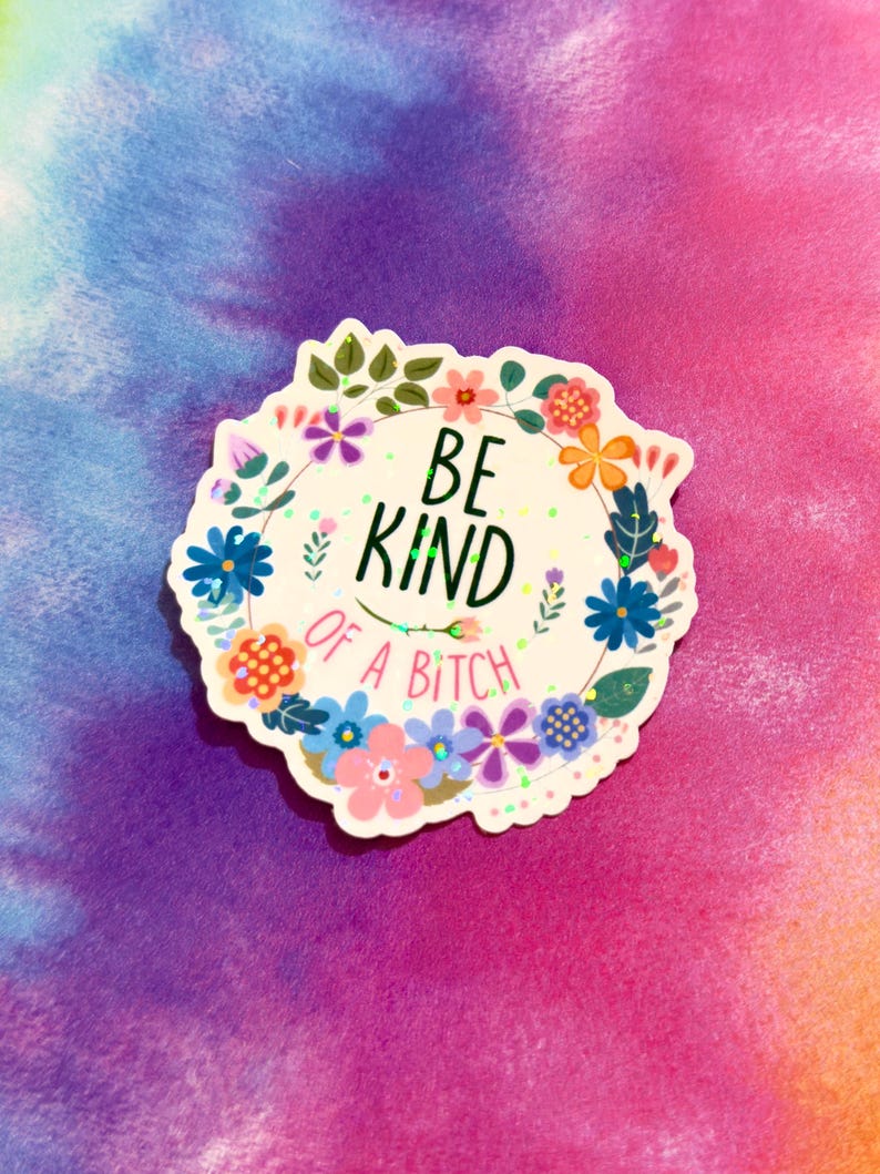 May include: A white sticker with a floral wreath design and the text "BE KIND OF A BITCH". The sticker features colorful flowers in shades of blue, pink, orange, and purple, with green leaves. The background is a tie-dye pattern.