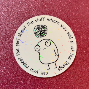 May include: A round white sticker with a cartoon figure and the text "can you repeat the part about the stuff where you said all of the things". The sticker has a holographic effect and is on a red surface.