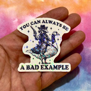May include: A die-cut sticker with a holographic finish featuring a black and white illustration of a raccoon wearing a cowboy hat and holding a pistol while riding a goose. The text reads YOU CAN ALWAYS BE A BAD EXAMPLE in dark blue letters.