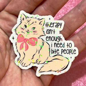 May include: A sticker featuring a cartoon cat with a pink bow and green eyes. The cat is surrounded by green glitter and the text reads "therapy isn't enough I need to bite people."