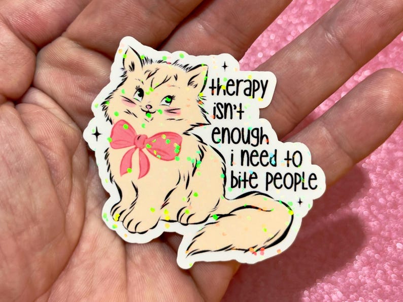 May include: A sticker featuring a cartoon cat with a pink bow and green eyes. The cat is surrounded by green glitter and the text reads "therapy isn't enough I need to bite people."