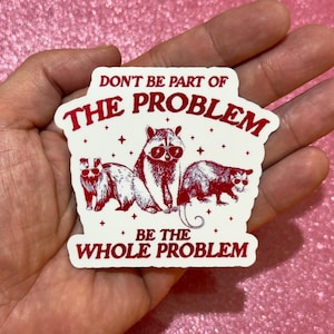 May include: A white sticker with red text and illustrations. The text reads "DON'T BE PART OF THE PROBLEM BE THE WHOLE PROBLEM." The sticker features three raccoons wearing sunglasses.